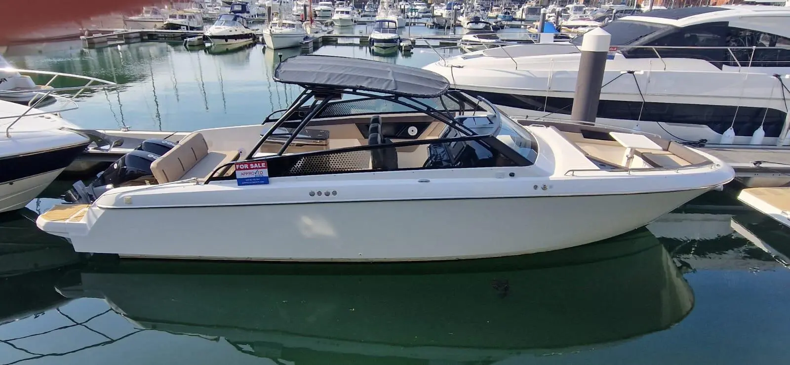 A white boat with a canopy docked at a marina, featuring 'FOR SALE' signage and visible reflections in the water.